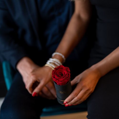 Prezent urodzinowy oraz na Walentynki Dzień Kobiet kwiaty mini flower box czerwona impregnowana wieczna róża w białym pudełku ze złotym logo Infinity Rose miłość para mężczyzna kobieta blondynka czarna sukienka perły bransoletka czerwony lakier do paznokci