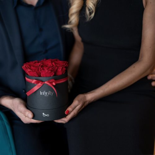 Prezent na urodziny Walentynki Dzień Kobiet kwiaty duży flowerbox czerwone stabilizowane wieczne róże w białym pudełku z srebrnym logo Infinity Rose czerwona wstążka para mężczyzna kobieta blondynka mała czarna sukienka